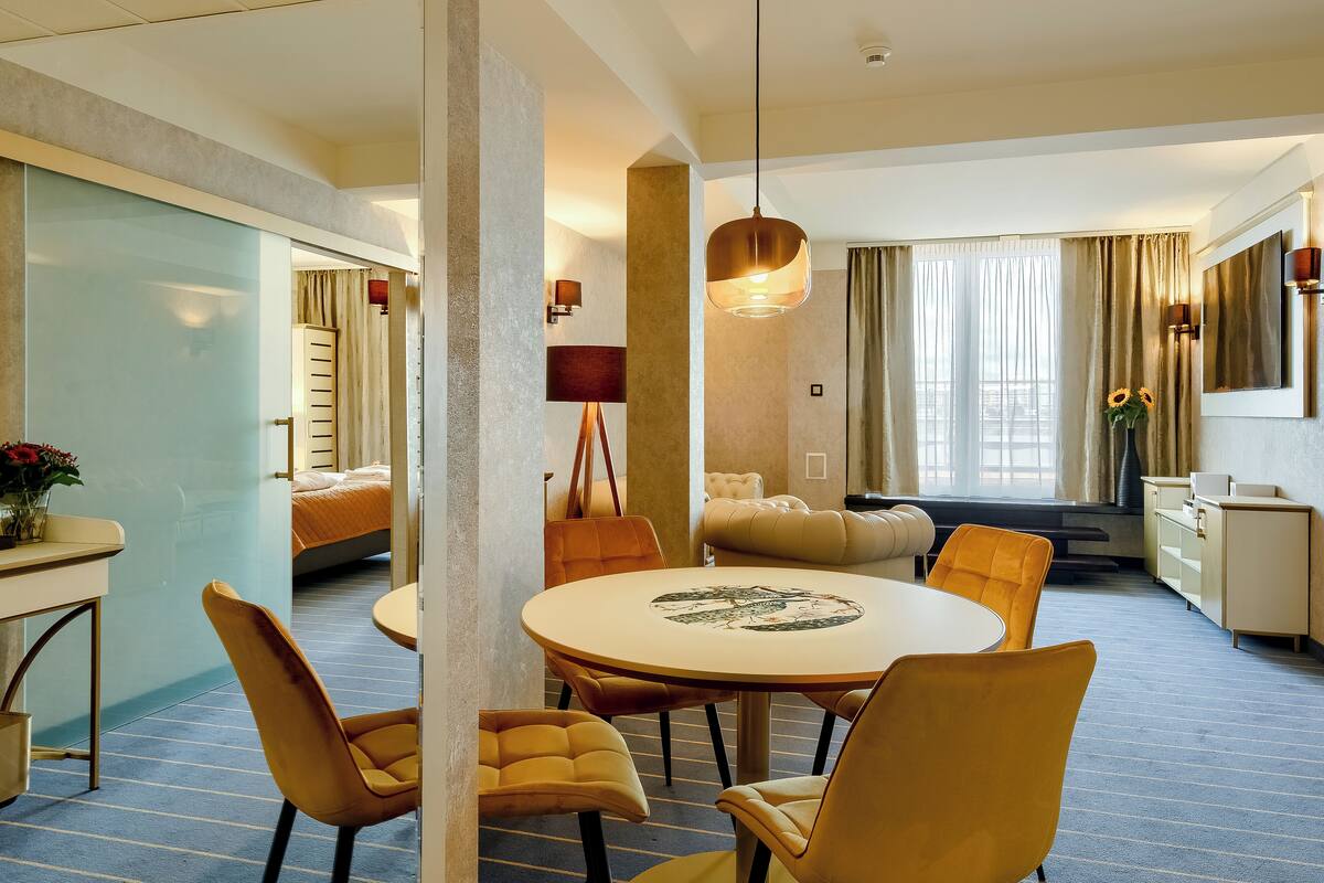 Een hotelkamer met een ronde eettafel, gele stoelen, een bank, een televisie en een groot raam met gordijnen.