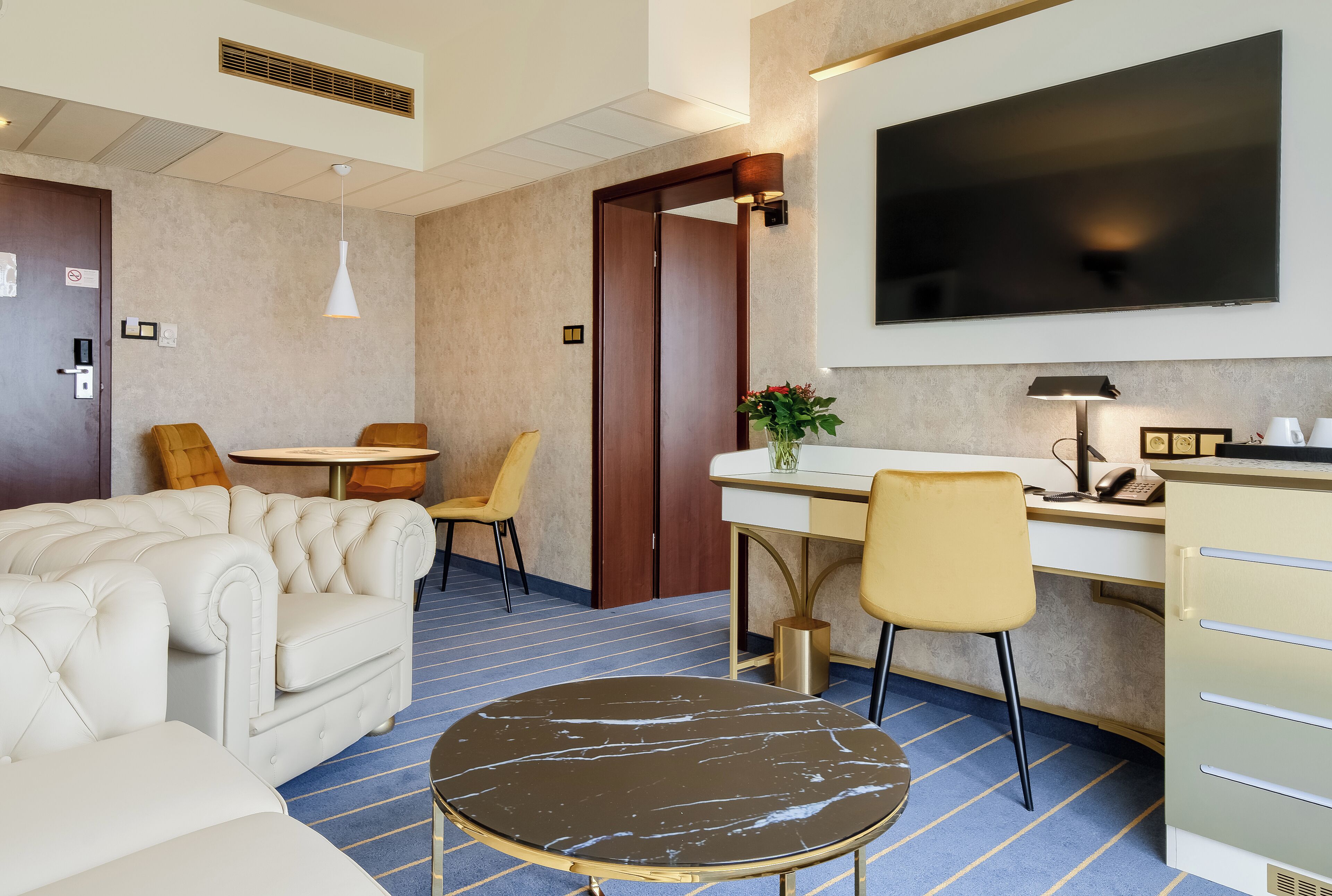 A hotel room with a sofa, a round table, a desk with a chair, a TV, and a minibar.
