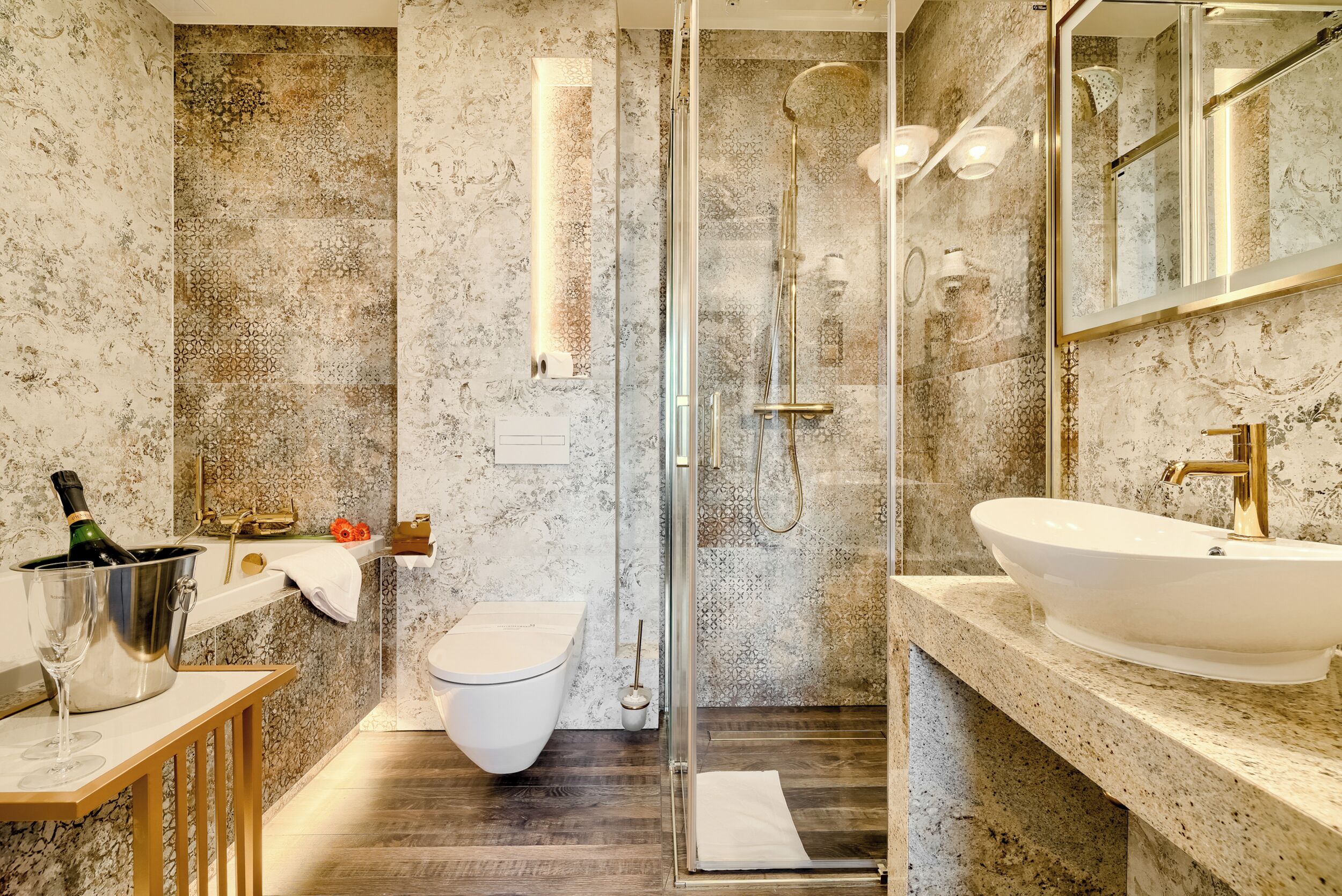 A bathroom with a marble wall, a white toilet, a glass-enclosed shower, a gold sink, and a wooden vanity.