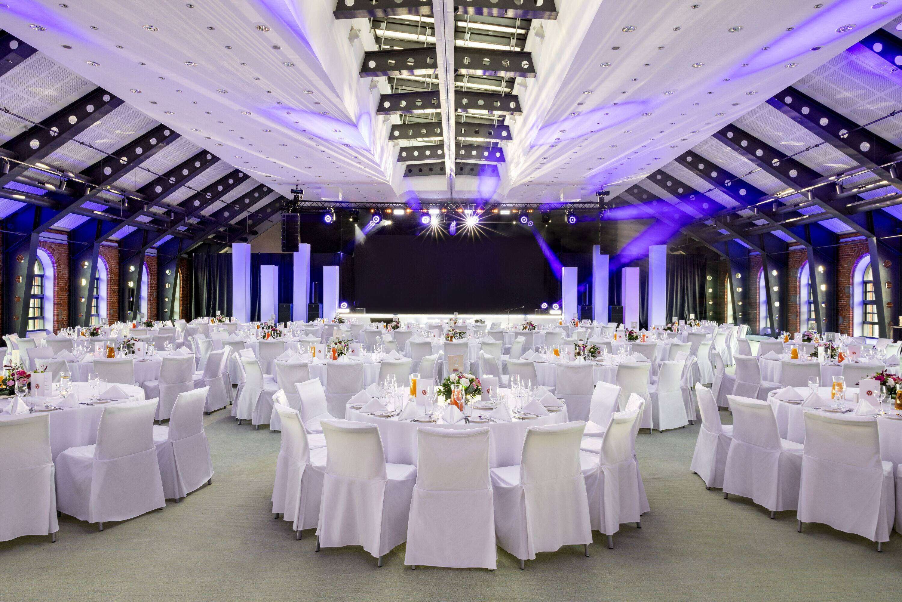 A large banquet hall with round tables and white tablecloths, featuring a high ceiling with exposed beams and a stage at the front.