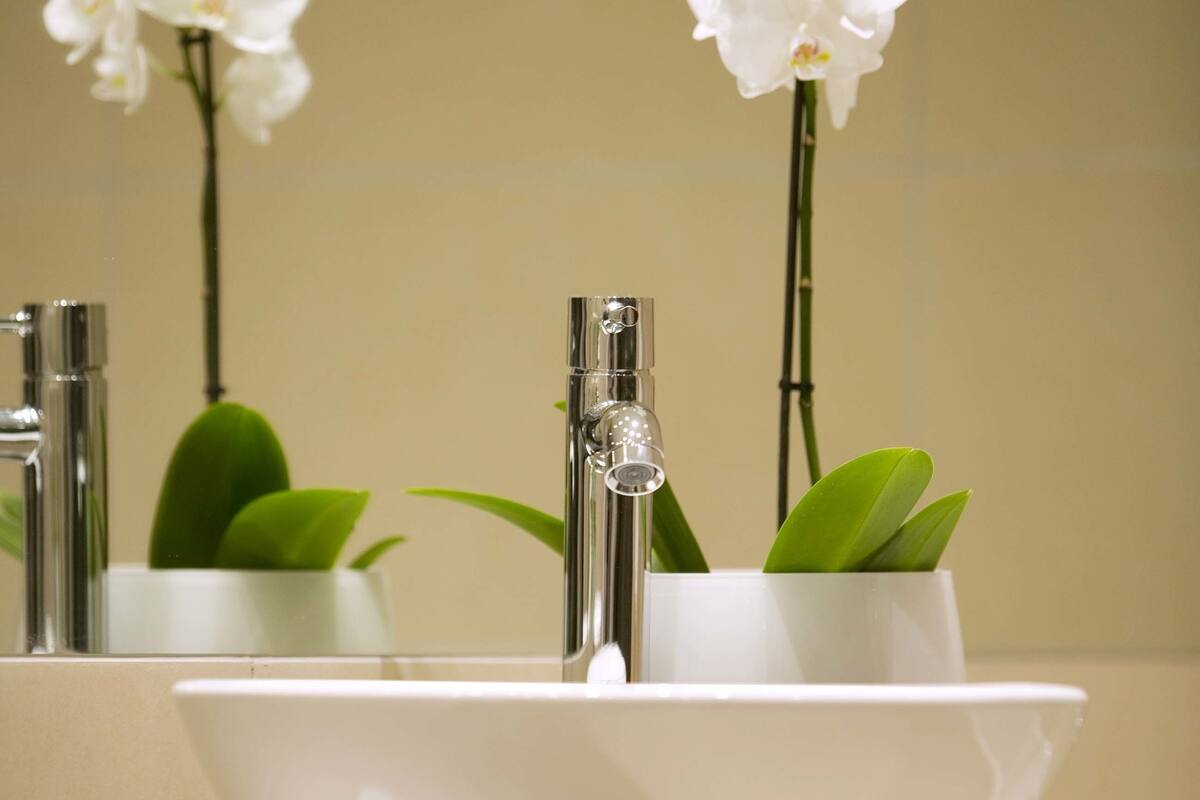 Un baño con un lavamanos blanco, dos llaves y un espejo que refleja orquídeas blancas.