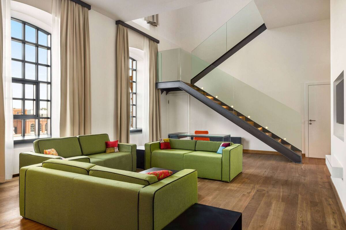 Un amplio salón con sofás verdes, una escalera y vistas a la ciudad a través de grandes ventanales.