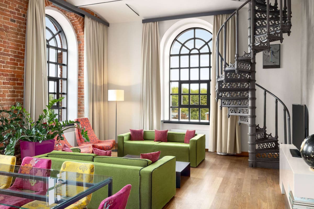 Amplia sala de estar con sofás verdes, una mesa de centro de vidrio y una pared de ladrillo. Se observa una escalera con barandillas ornamentales y un ventanal con cortinas.