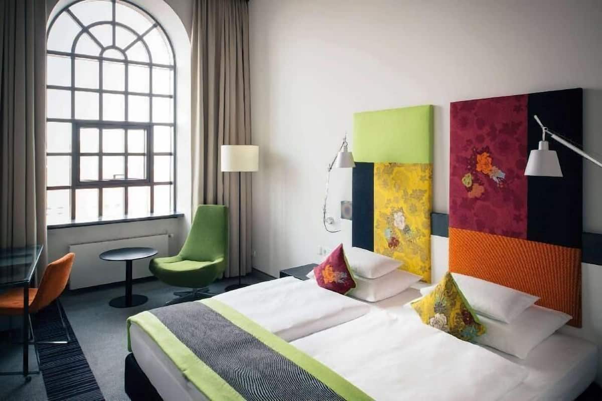 Habitación de hotel con cama, una silla, una mesita, una lámpara y ventana con cortinas.