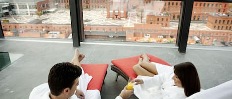 Una habitación de hotel moderna con ventanas amplias que ofrecen vista a la ciudad, un área de estar con cojines rojos y una mesa de vidrio.