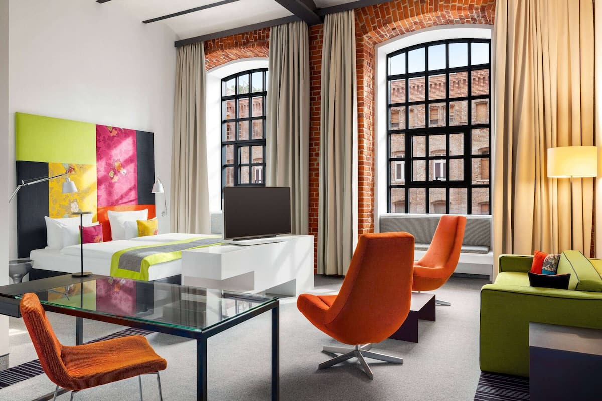 Una habitación de hotel moderna con una cama grande, un escritorio de vidrio, sillas naranjas, un sofá verde y un cuadro abstracto colorido.