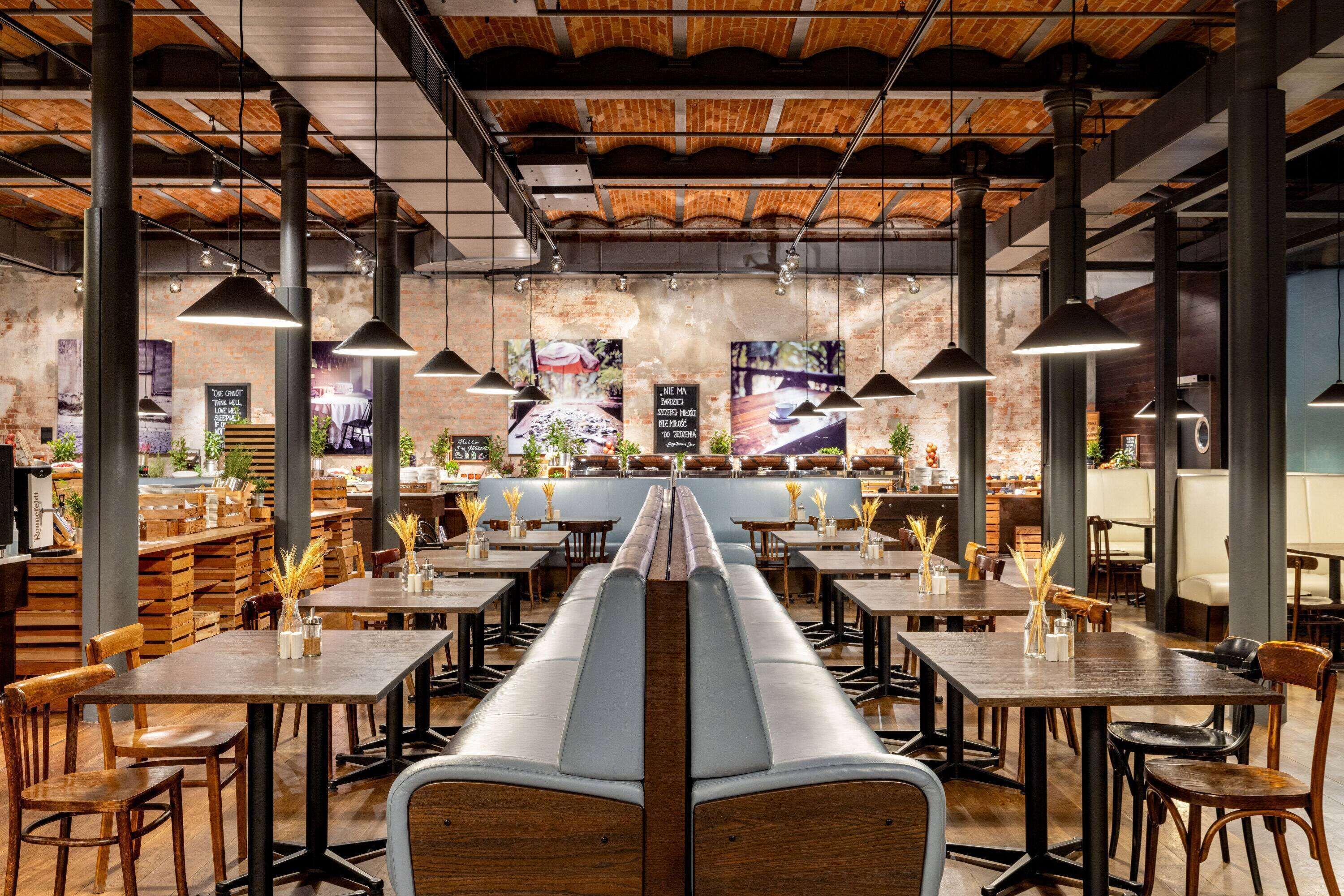 A restaurant with high wooden ceilings, exposed brick walls, and a central long bench. Tables are arranged around the bench, and pendant lights hang above.