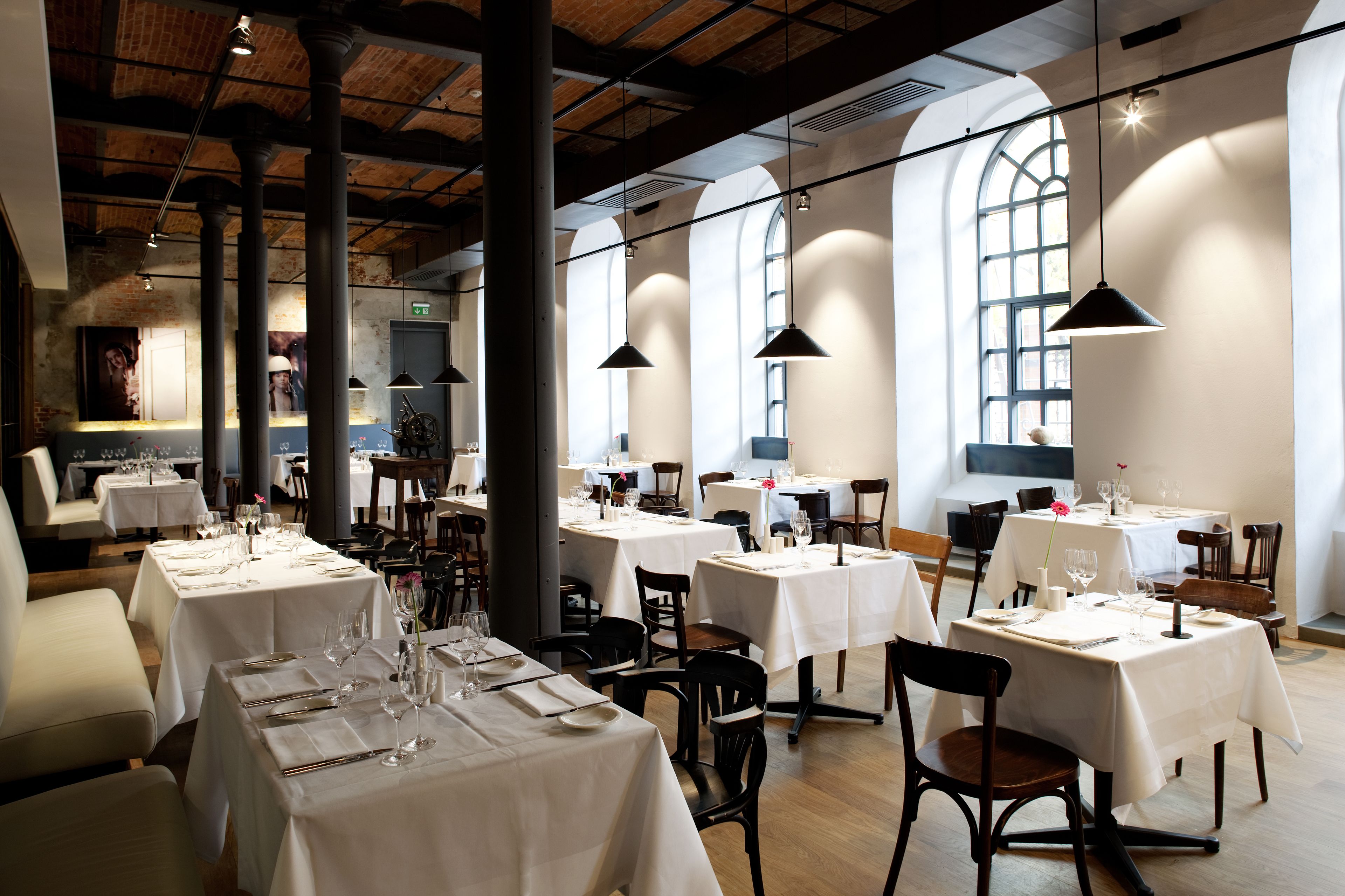 A restaurant with multiple tables set for dining, featuring a high ceiling with exposed beams and large windows allowing natural light.