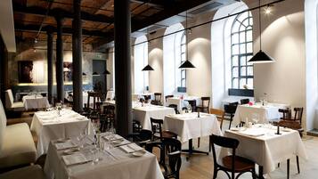 A restaurant with multiple tables set for dining, featuring a high ceiling with exposed beams and large windows allowing natural light.