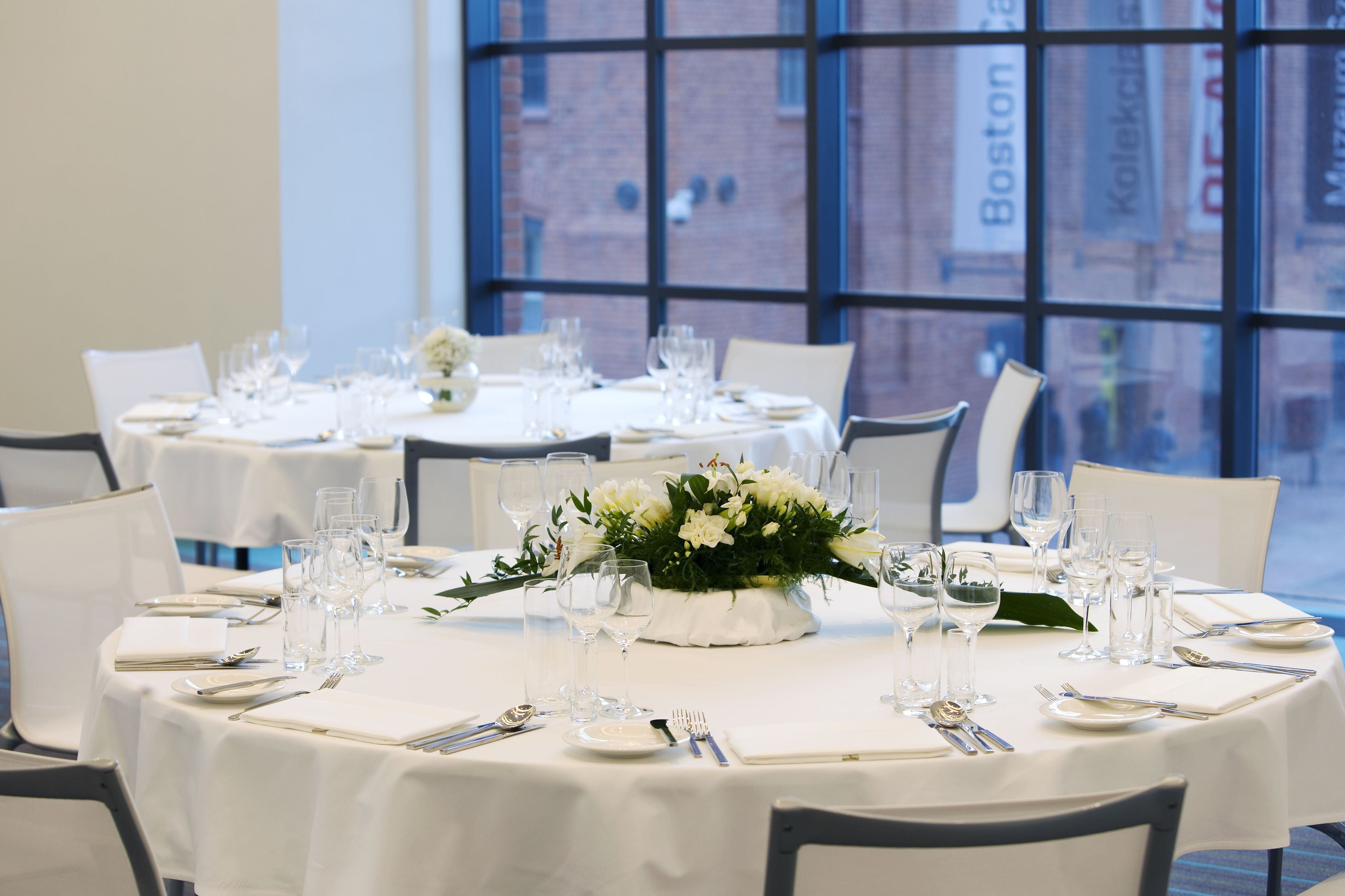 A dining area with round tables set for a meal, each with white tablecloths, glassware, and cutlery. A floral centerpiece adorns the tables. The room has large windows with a view of a brick building.
