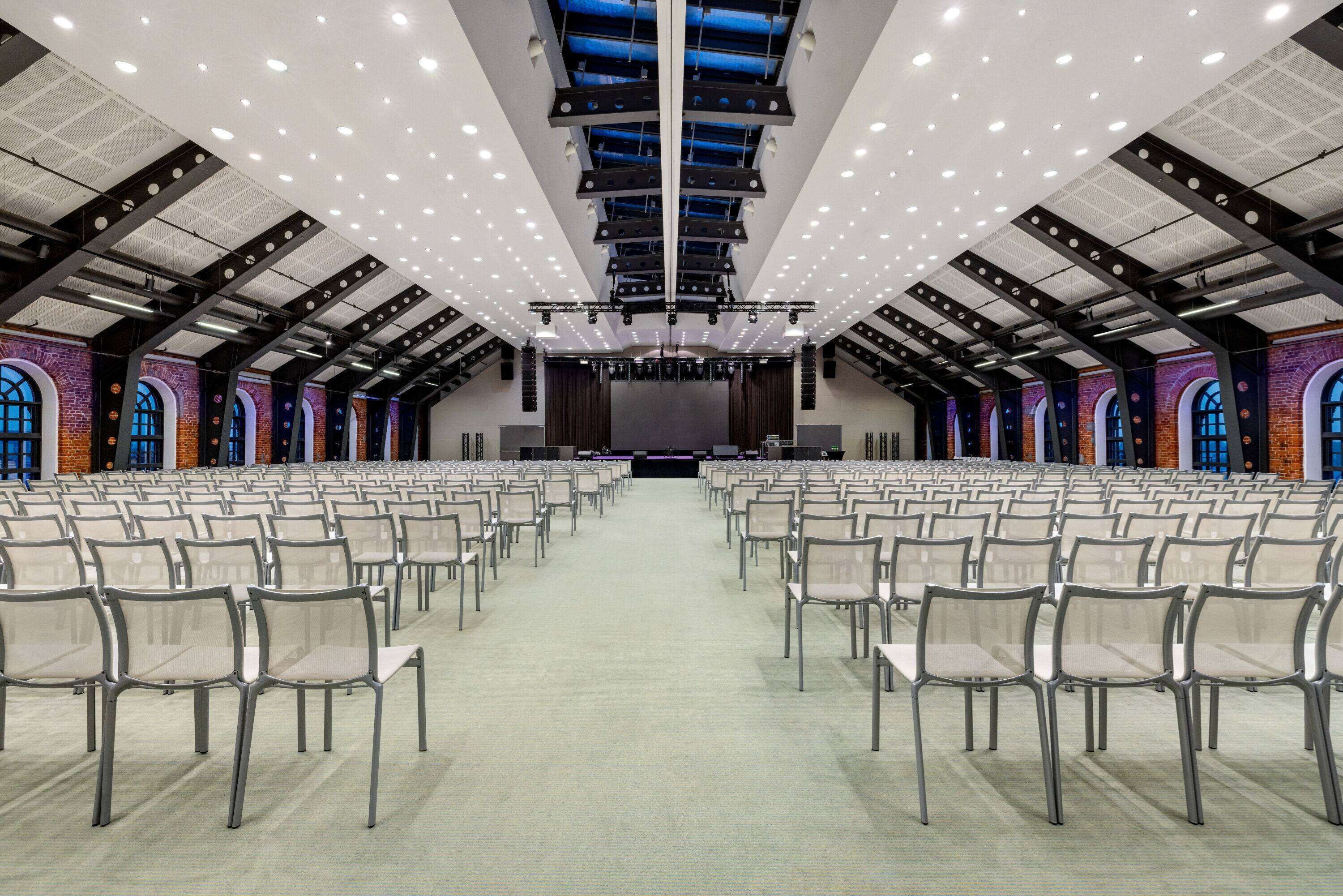 A large hall with rows of chairs facing a stage, high ceilings with exposed beams, and brick walls with arched windows.