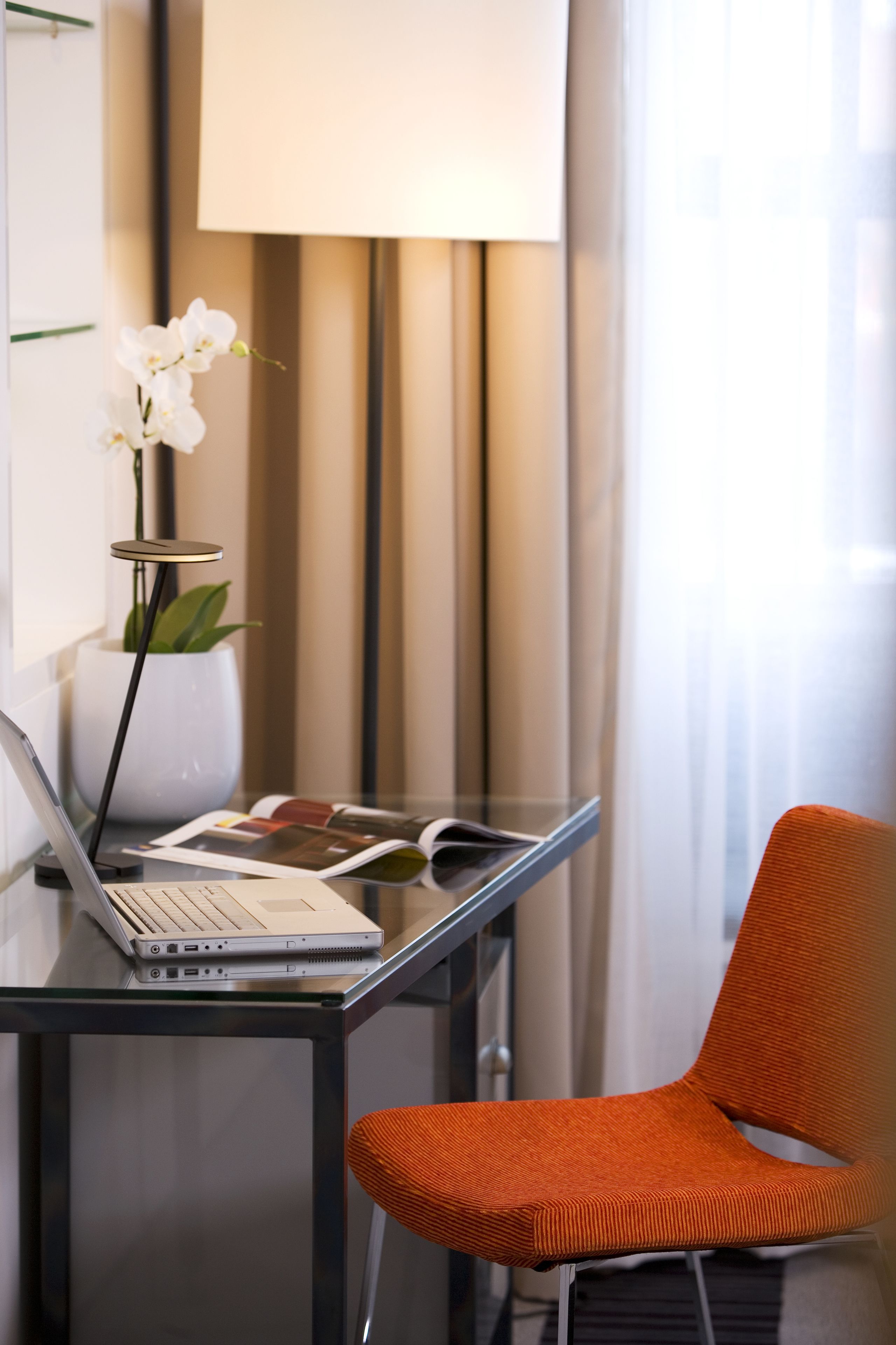 A modern hotel room with a glass desk, a laptop, a magazine, a potted orchid, and an orange chair.