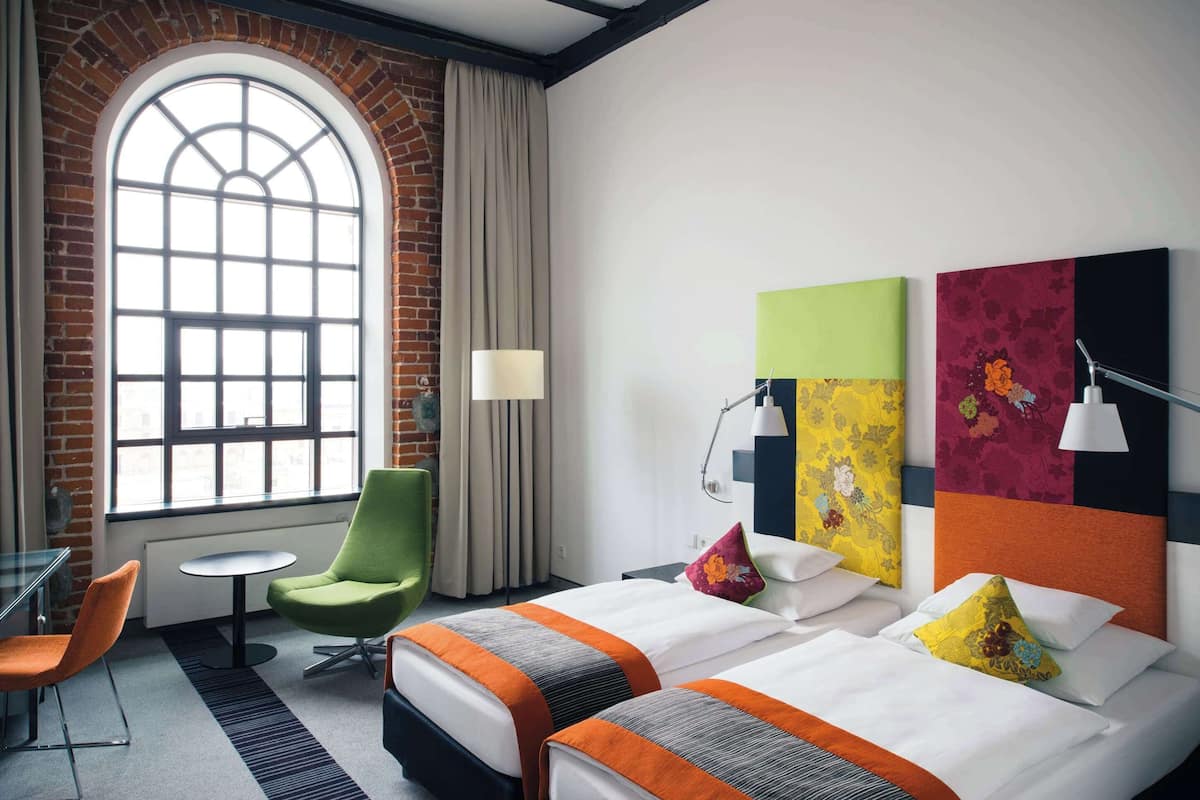 Habitación de hotel con dos camas, una silla verde, una mesa redonda y un amplio ventanal de arco.