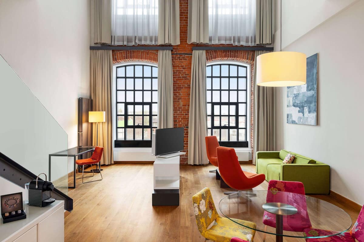 Una sala de estar moderna con una mesa de vidrio, sillas rojas y amarillas, un sofá verde y un televisor sobre un soporte blanco.
