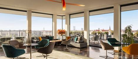 A spacious lounge area with modern furniture, large windows offering city views, and a high ceiling with orange accents.