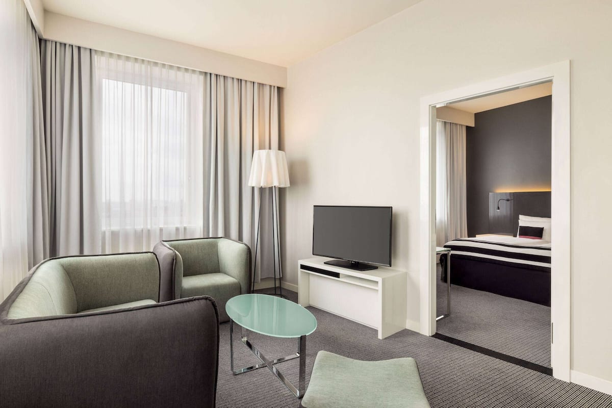 A hotel room with a sofa, a glass coffee table, a TV on a stand, a bed, and a desk.
