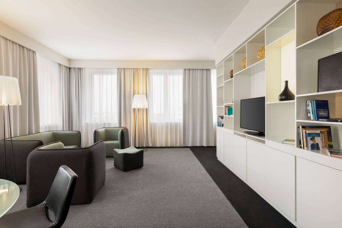 A modern hotel room with a sofa, armchairs, a TV, a bookshelf, and a large window with curtains.