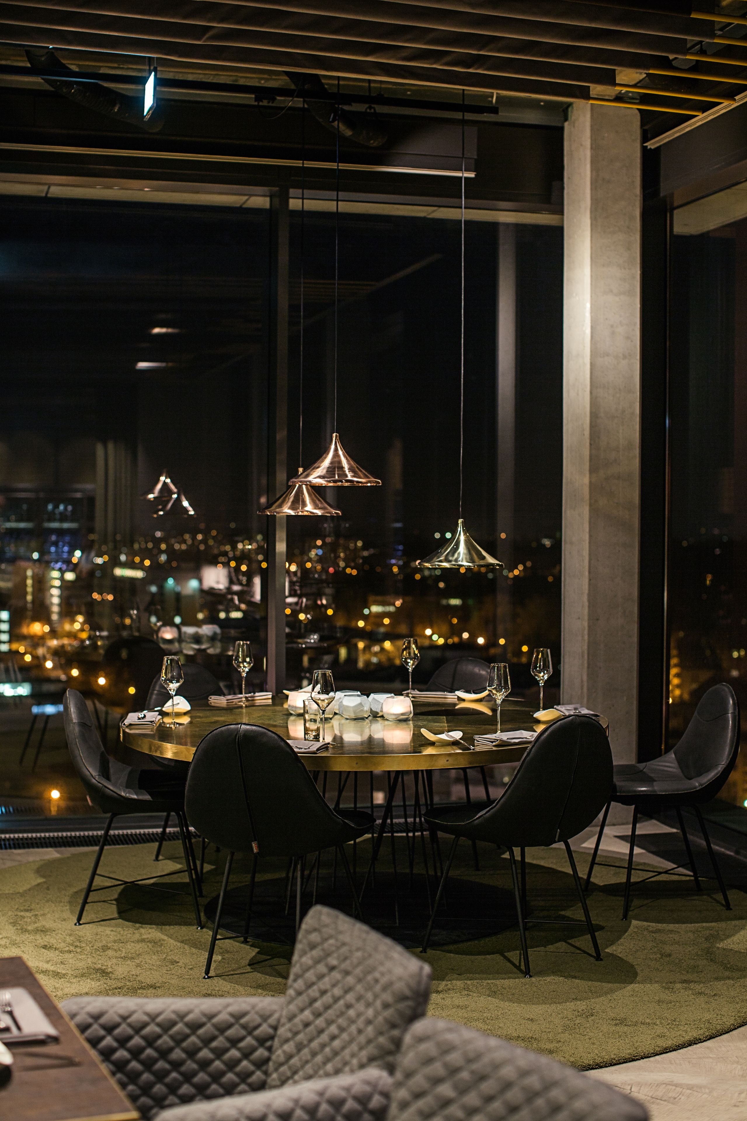 A dining area with a round table, black chairs, and pendant lighting. A cityscape view is visible through large windows.