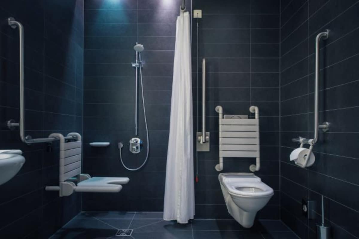Un baño moderno con ducha, inodoro y lavamanos, equipado con barras de apoyo y una cortina blanca.