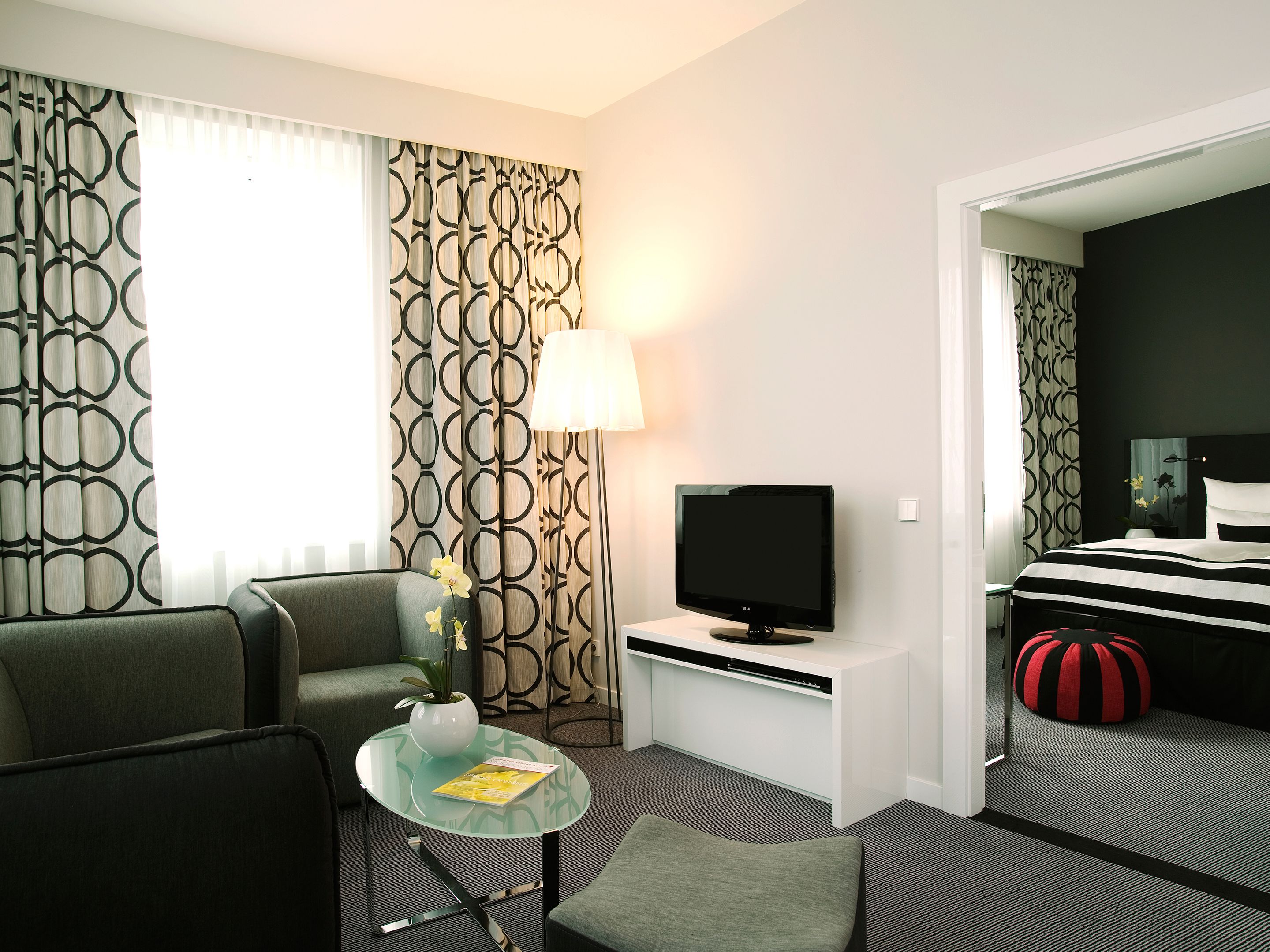 Ett hotellrum med en soffa, ett litet runt bord med en lampa, en tv och en säng med randigt sänglinne.