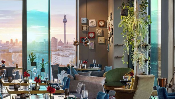 Un interior moderno con una vista panorámica de la ciudad, incluyendo una torre, comedor con mesas y sillas, y relojes decorativos y plantas.