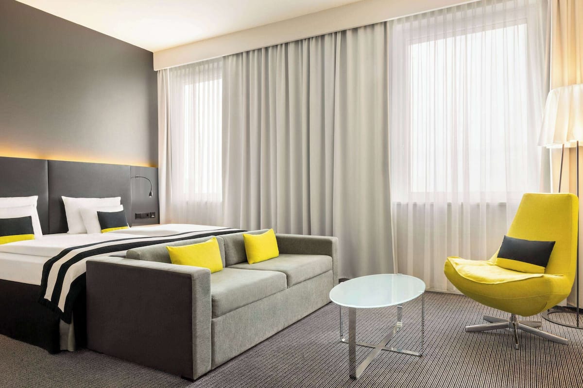 A hotel room with a bed, grey sofa, yellow armchair, and a small white table.
