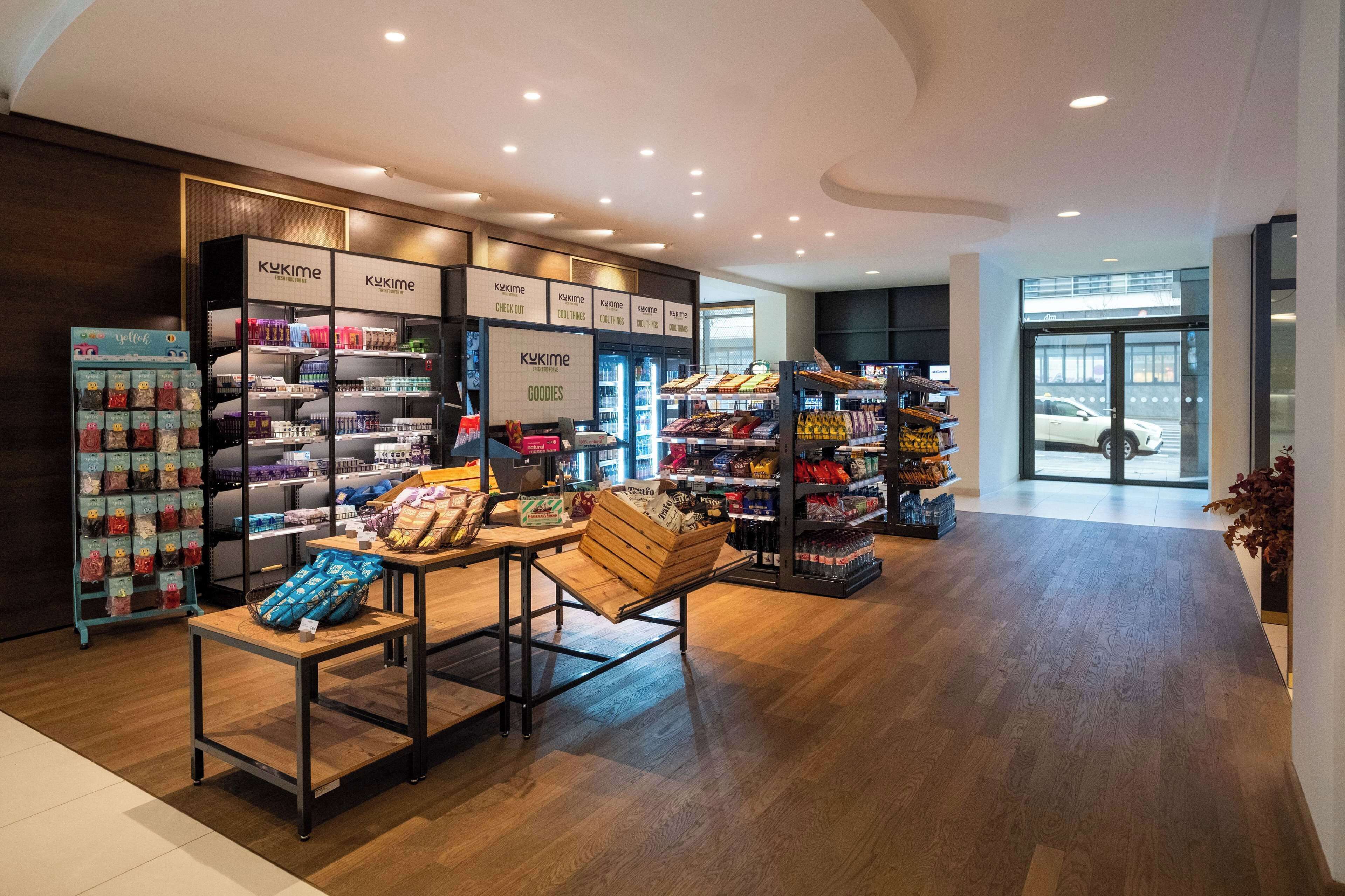 A modern retail store interior with shelves stocked with various products, a wooden display table, and a glass door entrance.