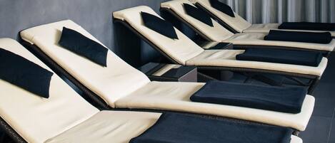 A row of sun loungers with cushions, arranged against a wall with a curtain.