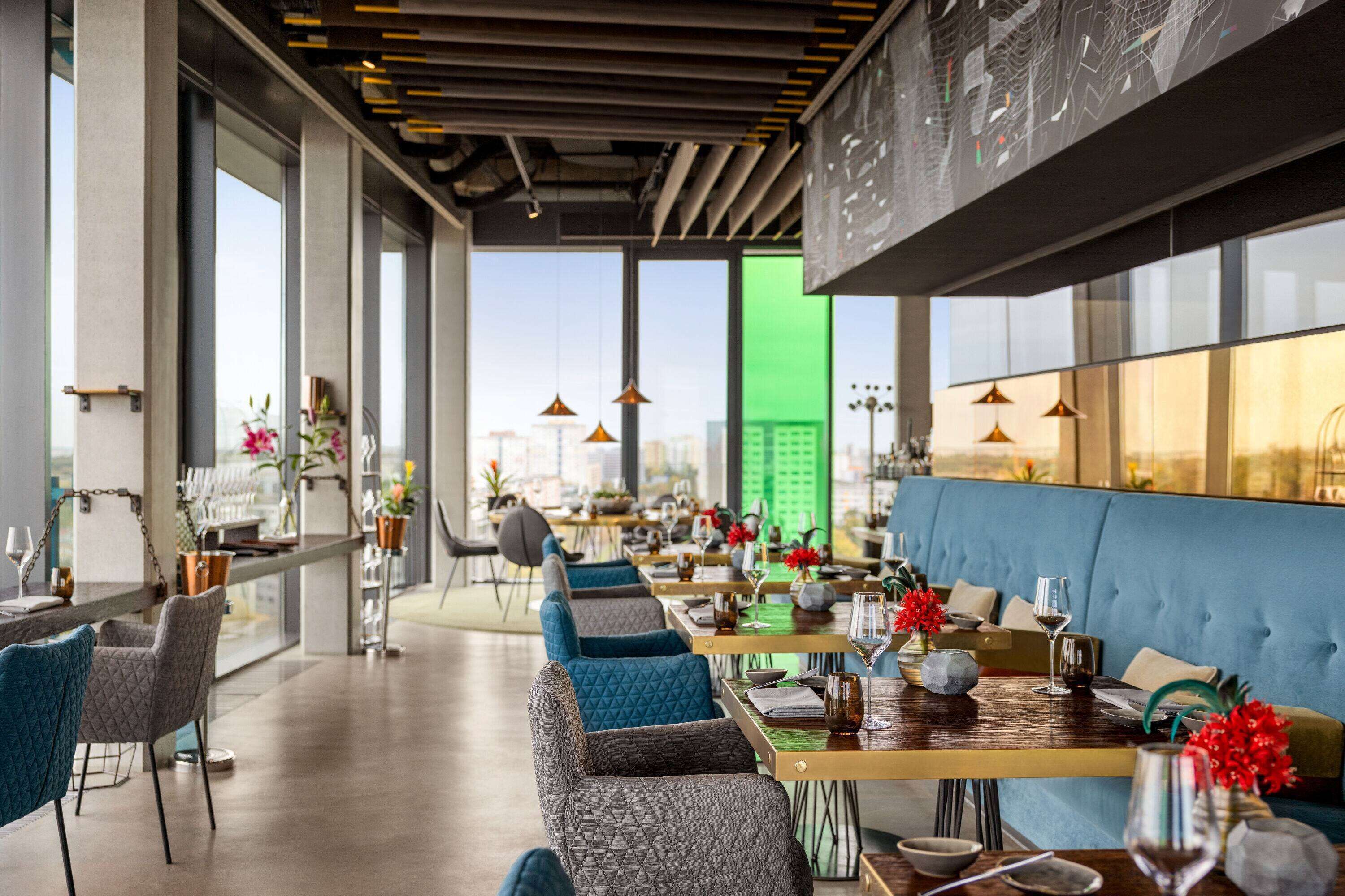 Un restaurante moderno con una barra alargada, asientos tapizados en azul y grandes ventanales con vistas a la ciudad.
