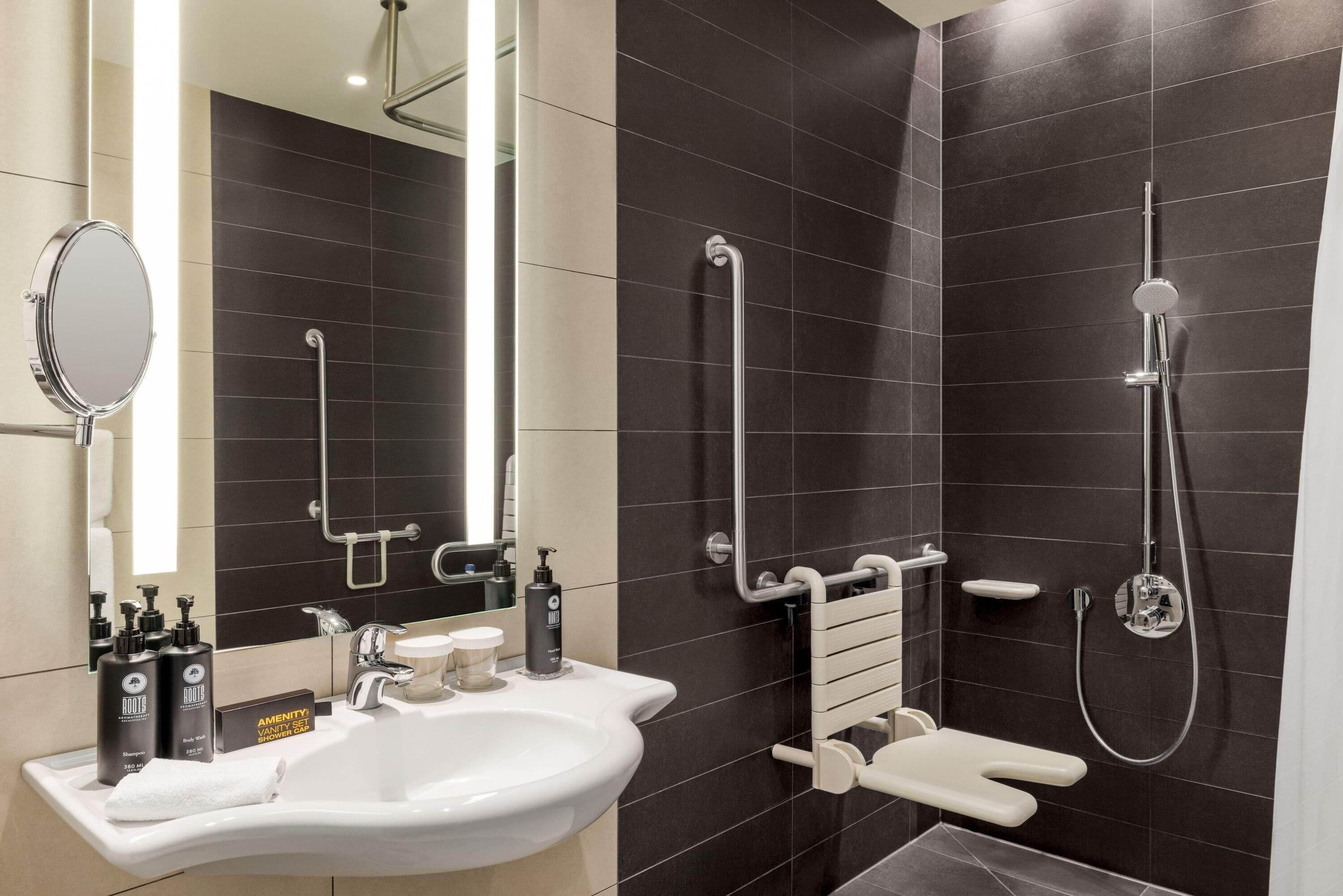 Un baño moderno con lavamanos blanco, espejo y paredes de azulejos oscuros. Incluye un cabezal de ducha manual y una barra de apoyo para mayor seguridad.