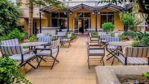 An outdoor patio with striped chairs and tables, surrounded by greenery and a building with large windows. - Achillion Palace Hotel Rethymno City Center (Rethymno)