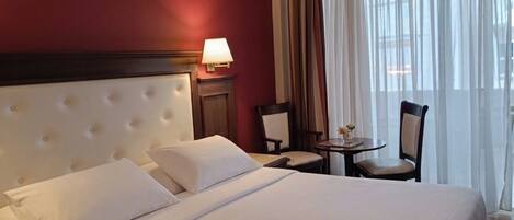 A hotel room with a large bed, a bedside table, a lamp, a chair, and a view of the outdoors through sheer curtains.