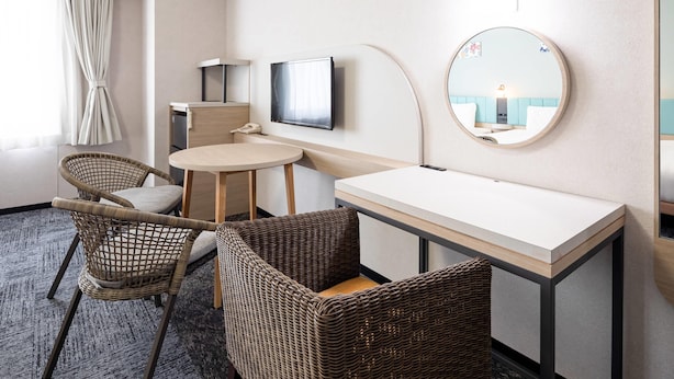 Une chambre d’hôtel avec un miroir rond, une télévision, une petite table, des chaises en osier et une fenêtre avec des rideaux.