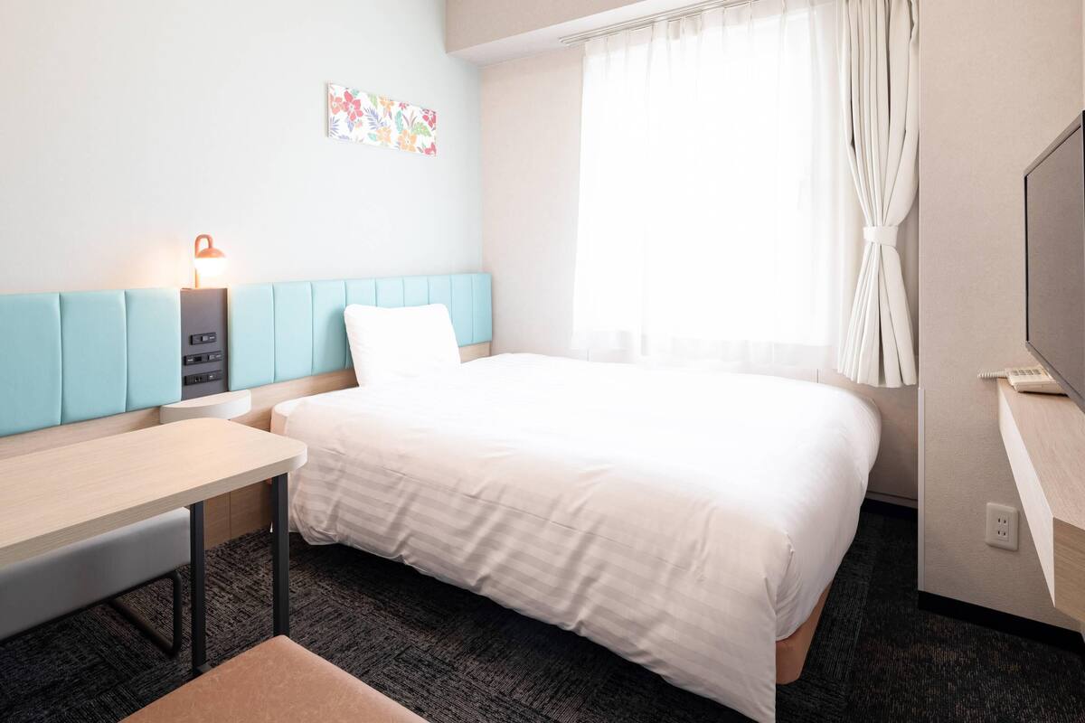 Quarto de hotel com cama, escrivaninha, cadeira, luminária, televisão e janela com cortinas.