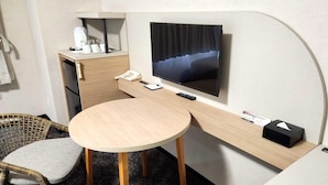 Une chambre d’hôtel avec un lit, un téléviseur fixé au mur, un petit bureau avec un téléphone, une télécommande et une boîte de mouchoirs. On y trouve également deux fauteuils en osier avec des coussins gris.