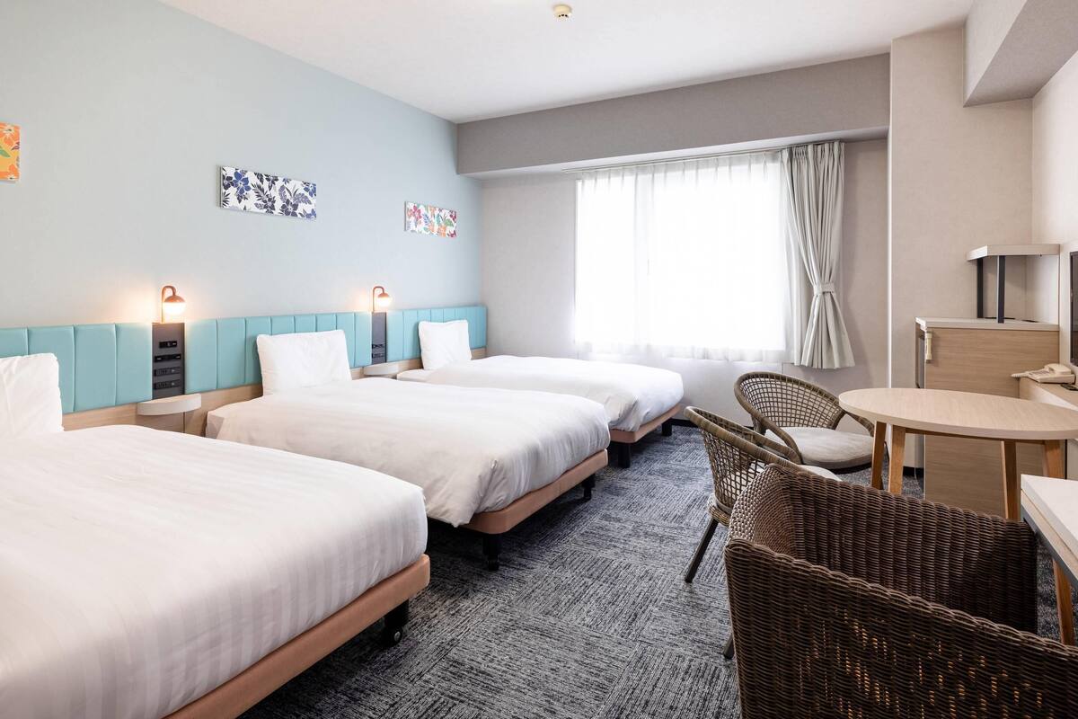 Quarto de hotel com três camas, uma escrivaninha, uma cadeira e uma janela grande com cortinas.