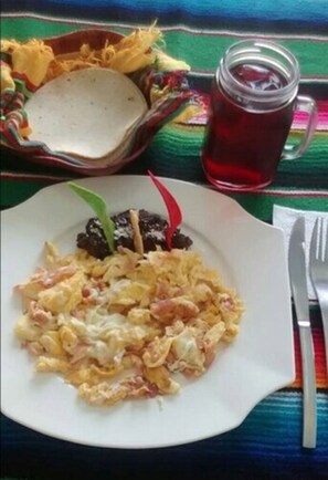 Un plato de huevos revueltos, un acompañamiento, un vaso de bebida roja y un mantel tejido de colores.