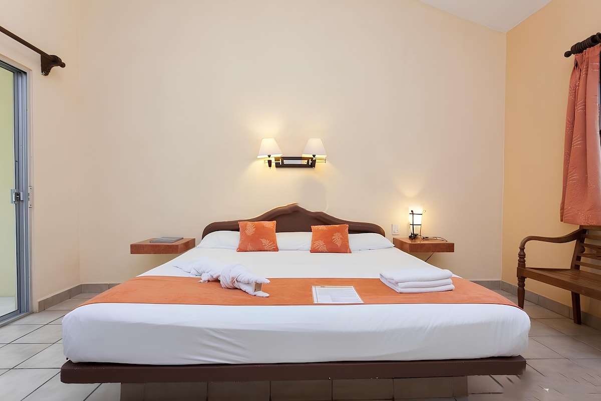 Una cama bien hecha con almohadas, una mesita de noche, una lámpara de pared y un banco.