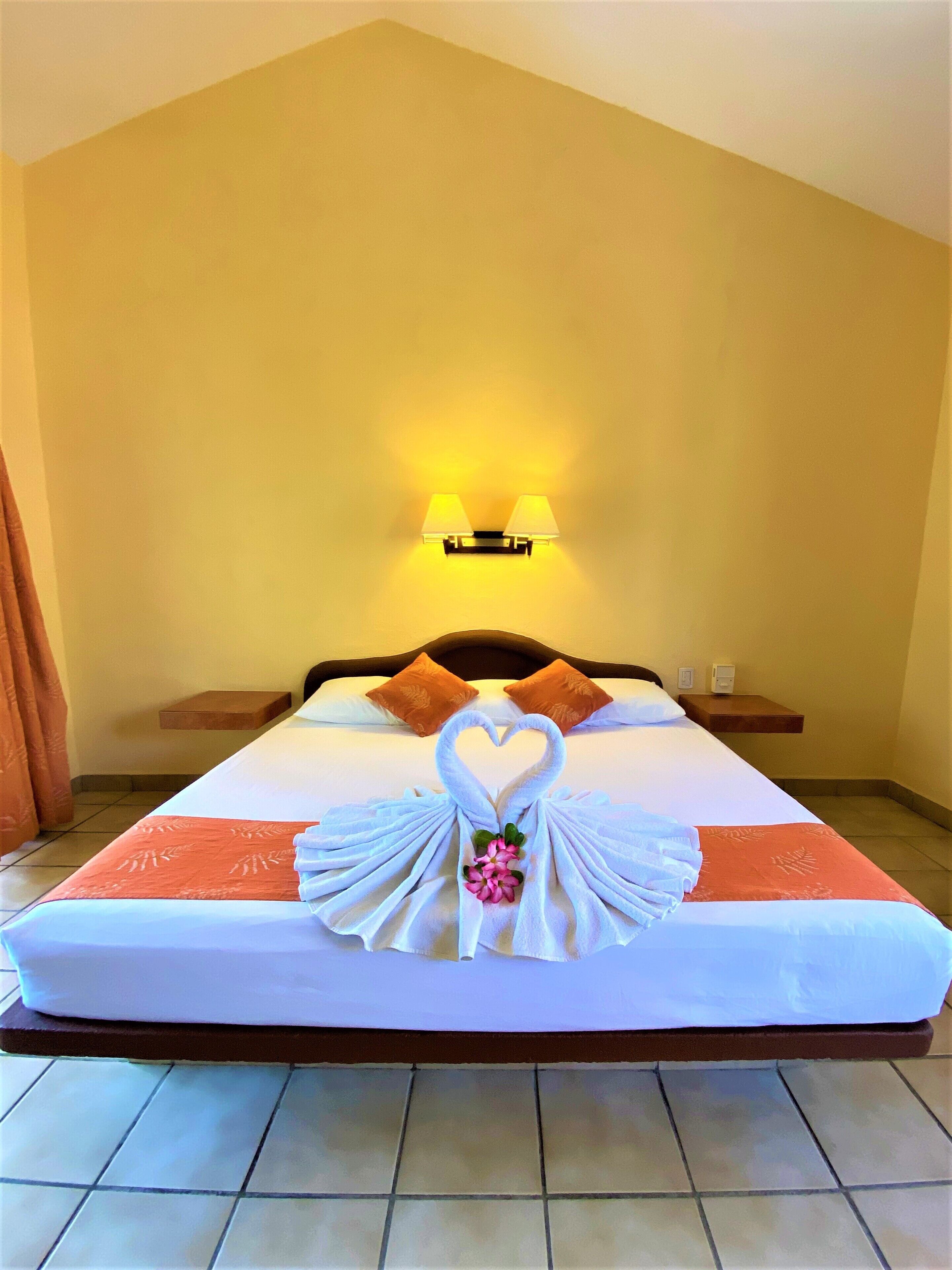 A hotel room with a bed, two pillows, a nightstand, a wall-mounted lamp, and a decorative towel arrangement on the bed.