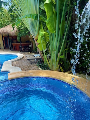 Una piscina con caída de agua, rodeada de exuberante vegetación tropical y una estructura de techo de paja.