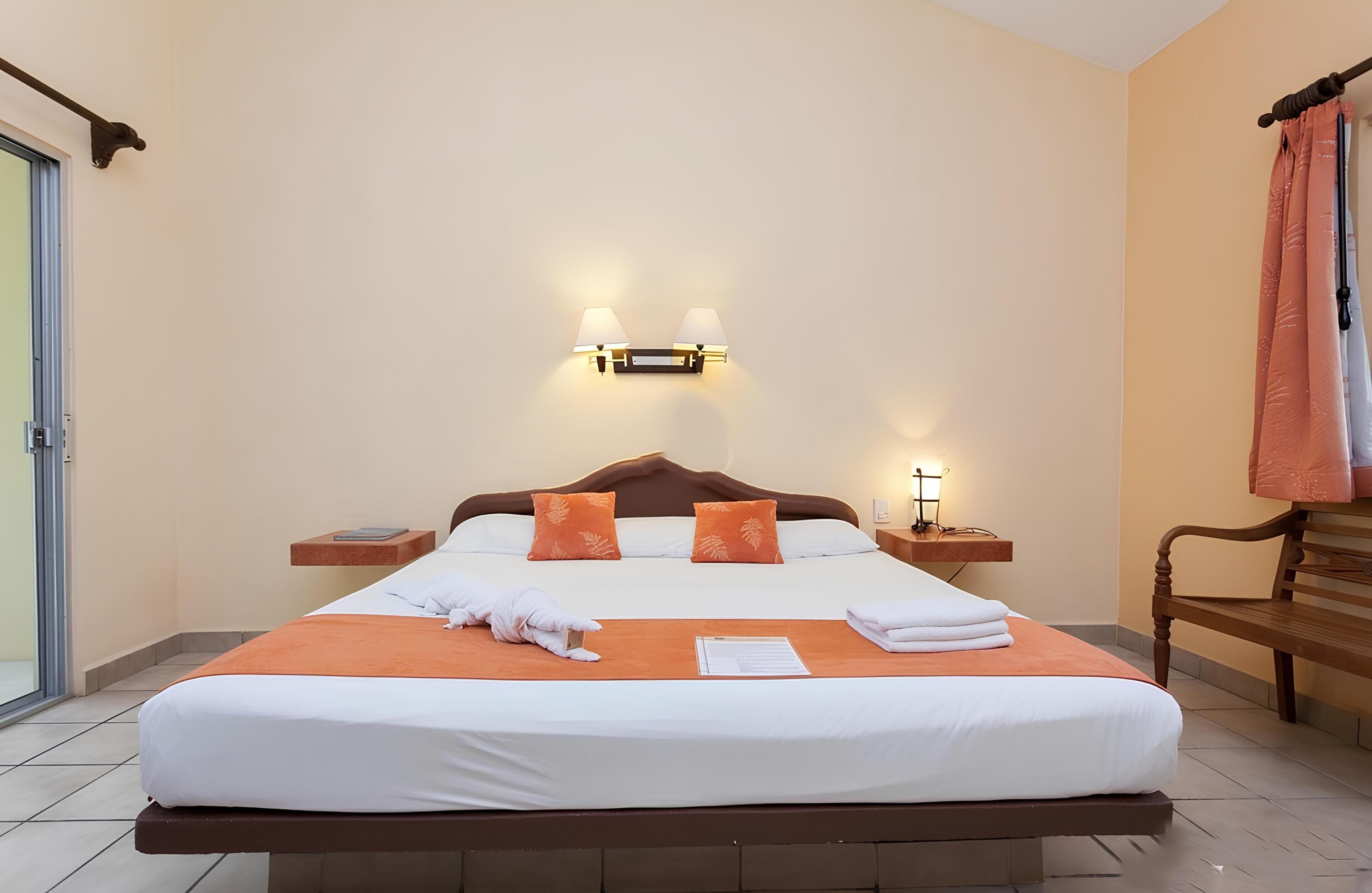 Een opgemaakt bed met kussens, een nachtkastje, een wandlamp en een bankje.