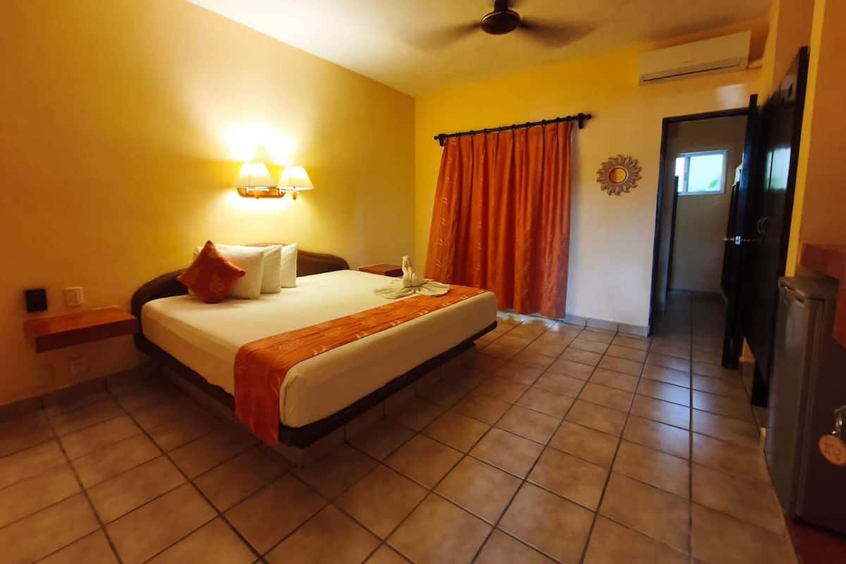 Una camera d'albergo con un letto, una ventola a soffitto, un condizionatore d'aria e una porta che dà su un corridoio.