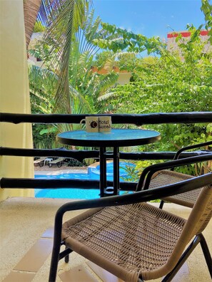 Un balcón con una silla de mimbre, una pequeña mesa con dos tazas y vistas a una piscina y exuberante vegetación.