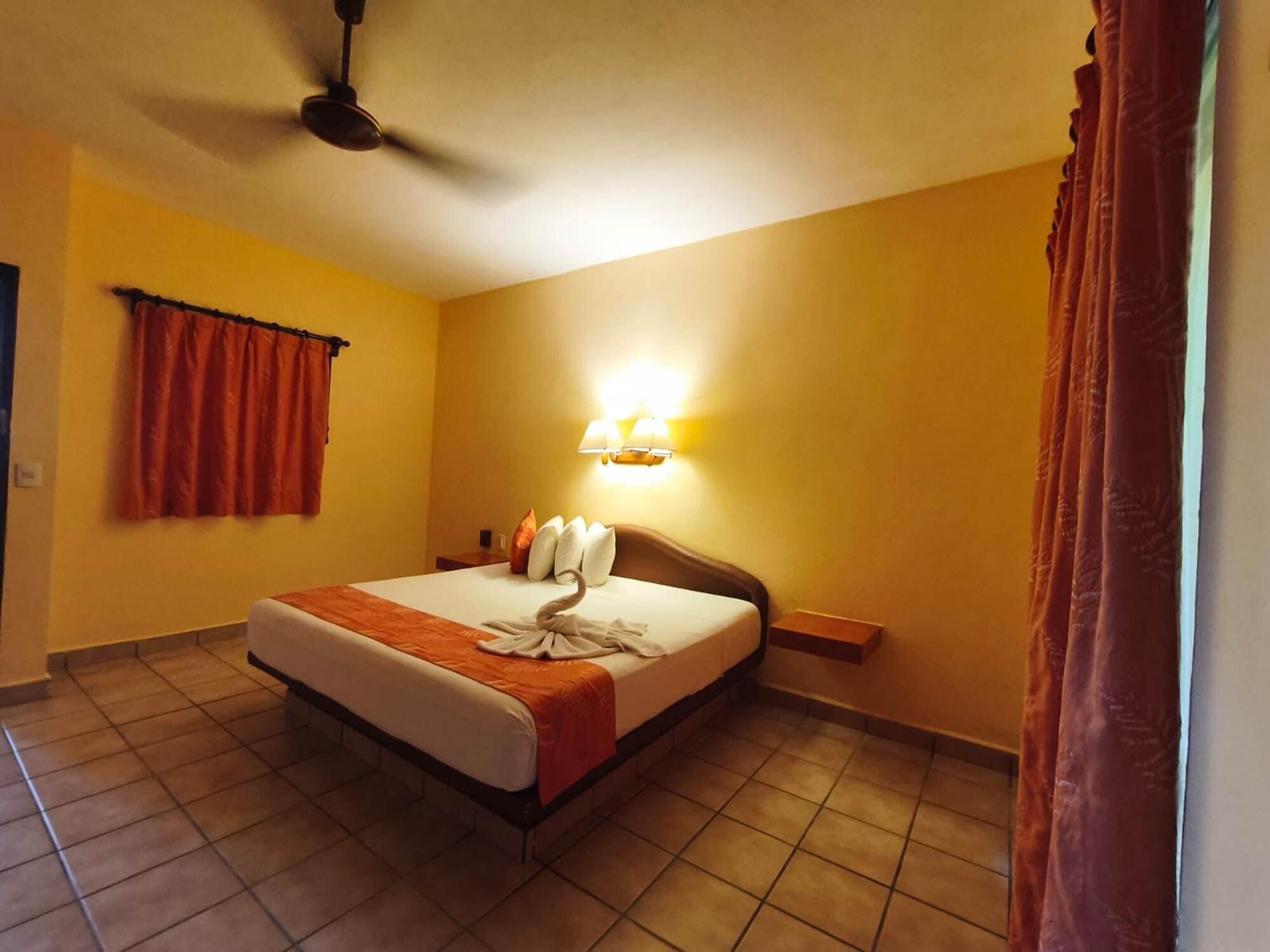 A hotel room with a bed, a ceiling fan, a wall-mounted lamp, and a window with curtains.