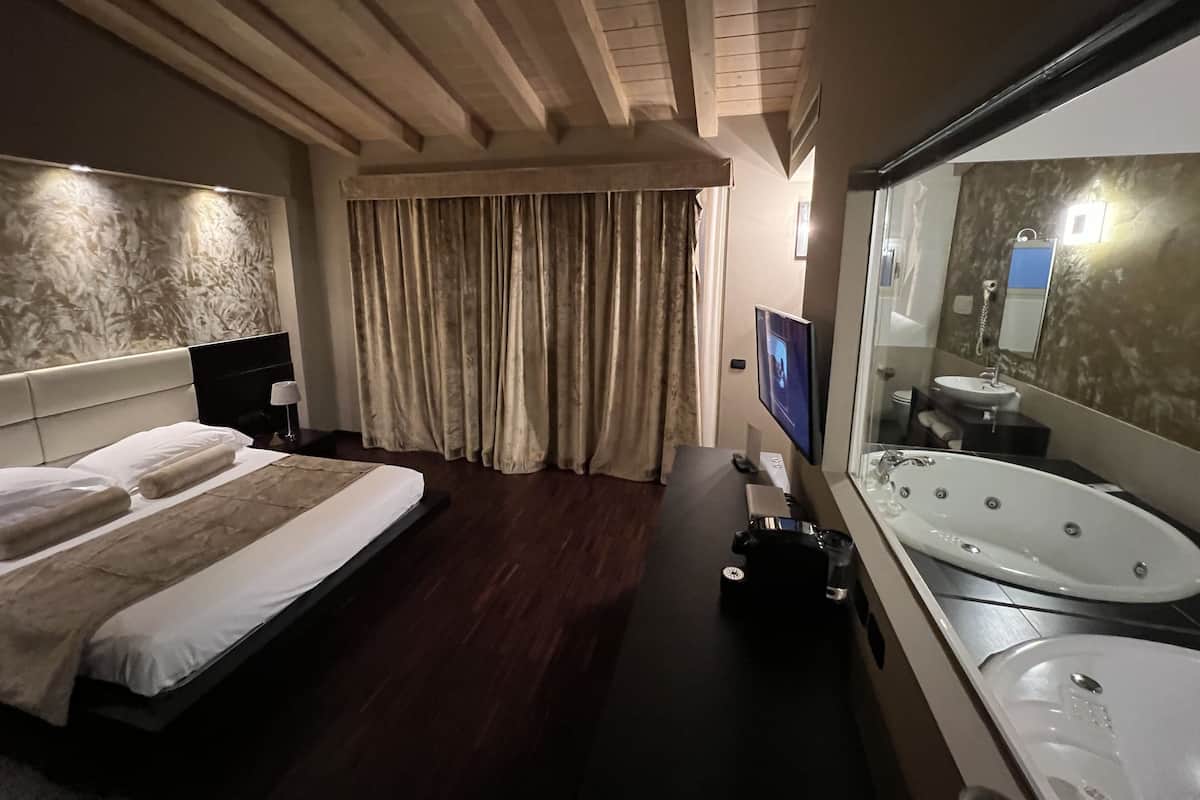 A hotel room with a large bed, a TV, a bathtub, and a mirror.