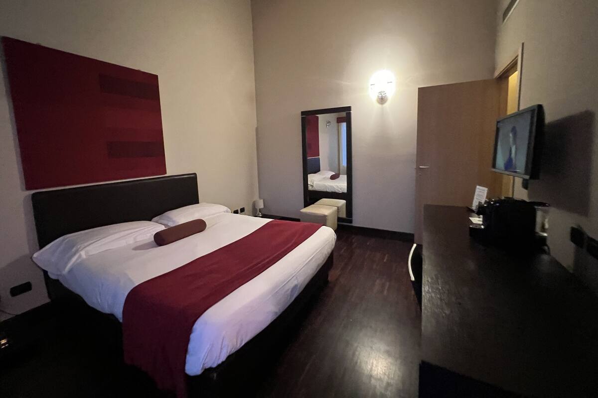 A hotel room with a large bed, a bedside table, a mirror, a TV, and a red accent wall.