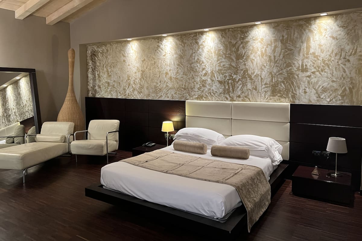 A modern hotel room with a large bed, a sitting area with two chairs, a large vase, and a wall-mounted lamp.
