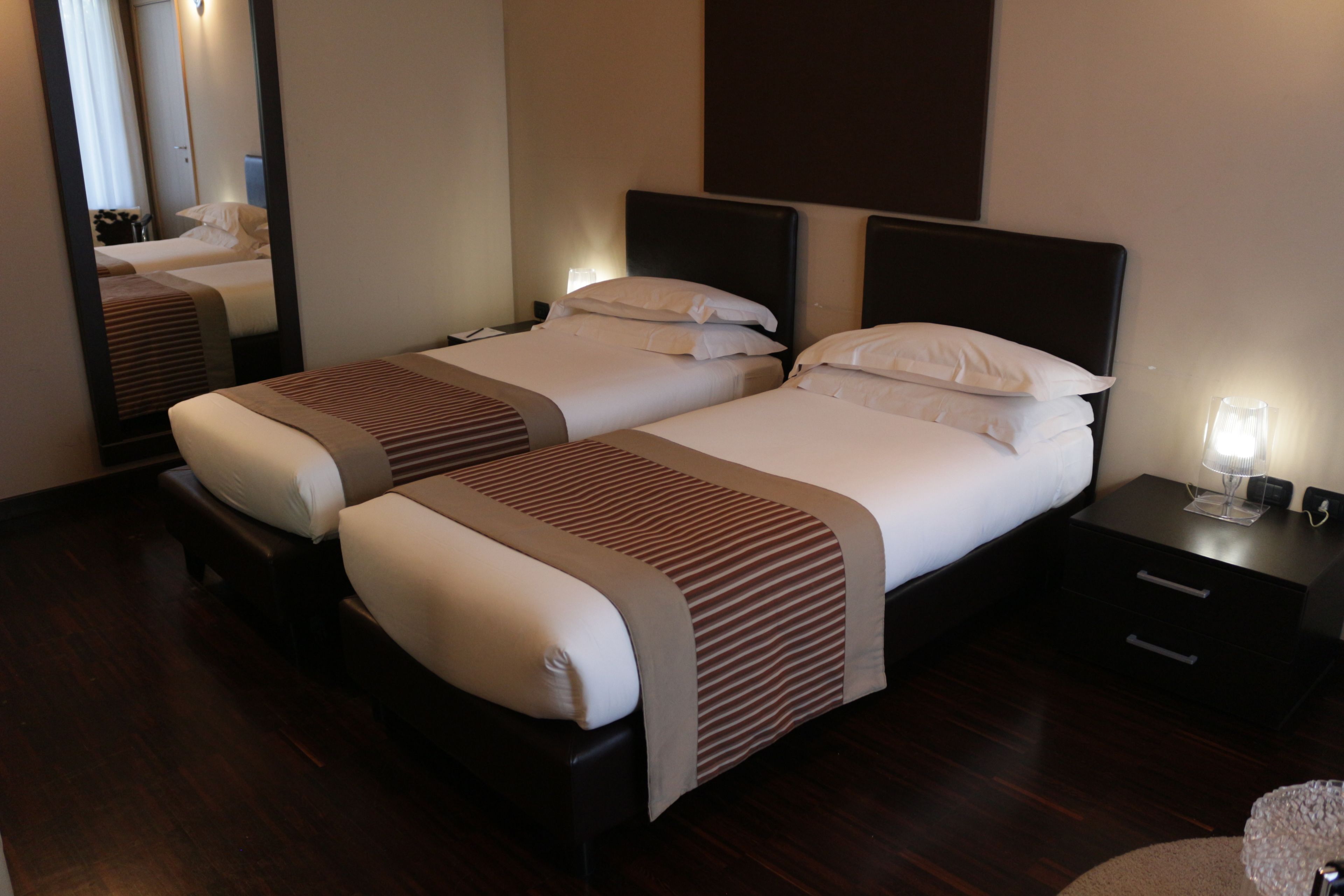 A hotel room with two beds, dark wooden flooring, and a nightstand with a lamp.
