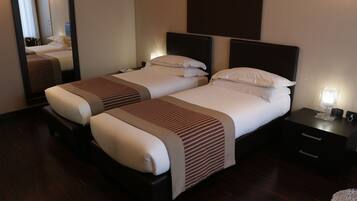 A hotel room with two beds, dark wooden flooring, and a nightstand with a lamp.