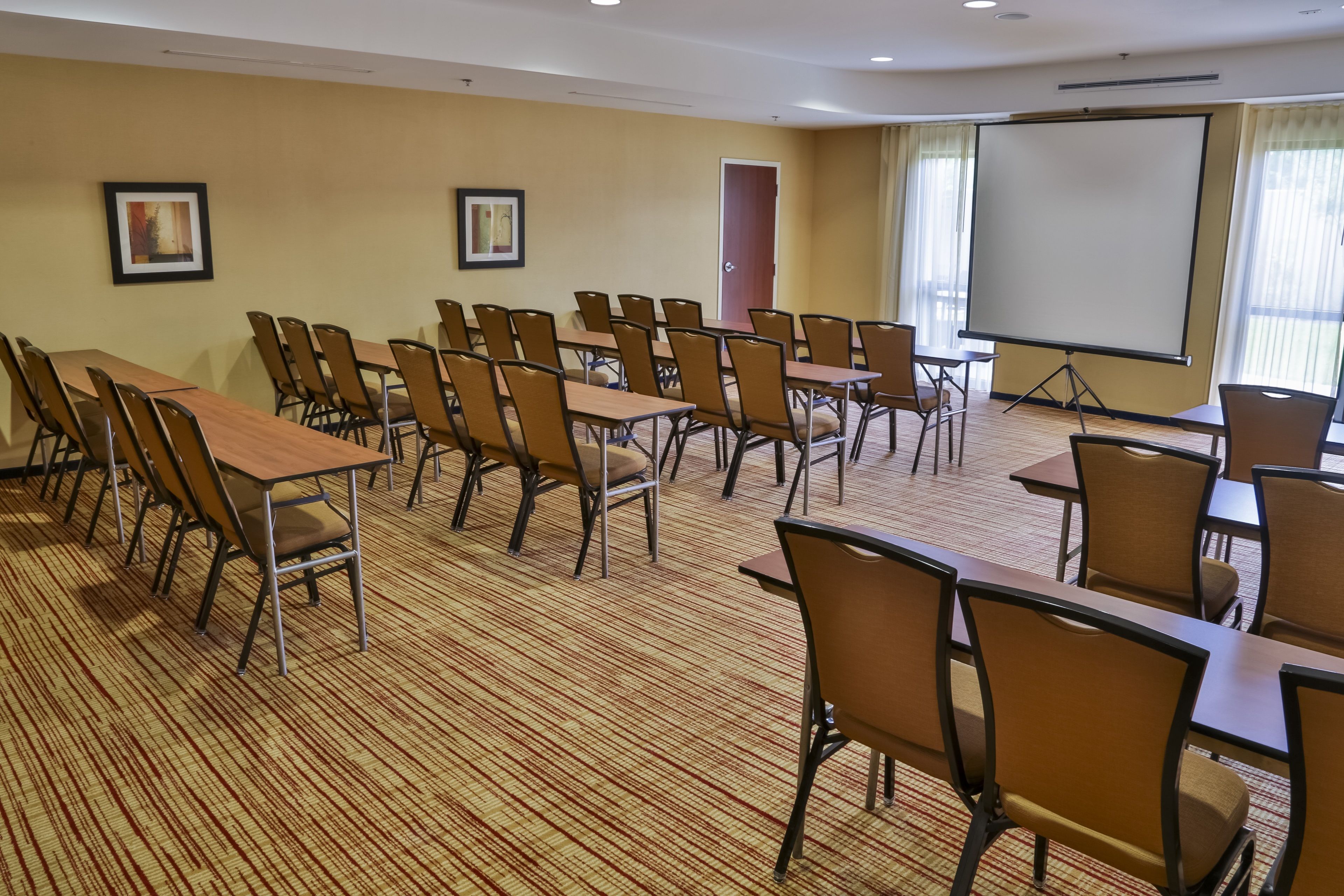 A conference room with multiple tables and chairs, a projector screen, and framed pictures on the walls.